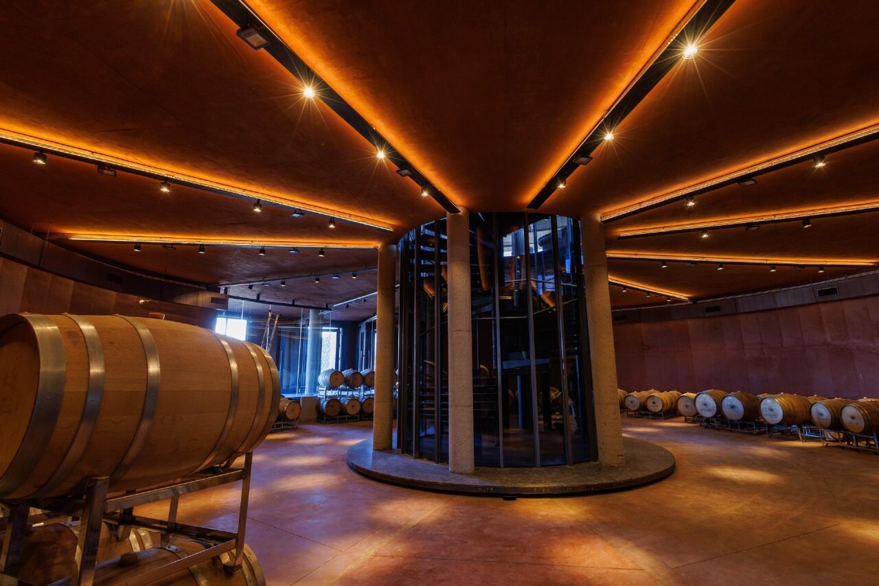 Barrel room lit in amber, oak barrels along both walls and a glassed-in tank at the centre